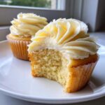 Close-up of two fluffy vanilla cupcakes, one with a bite revealing the moist crumb.