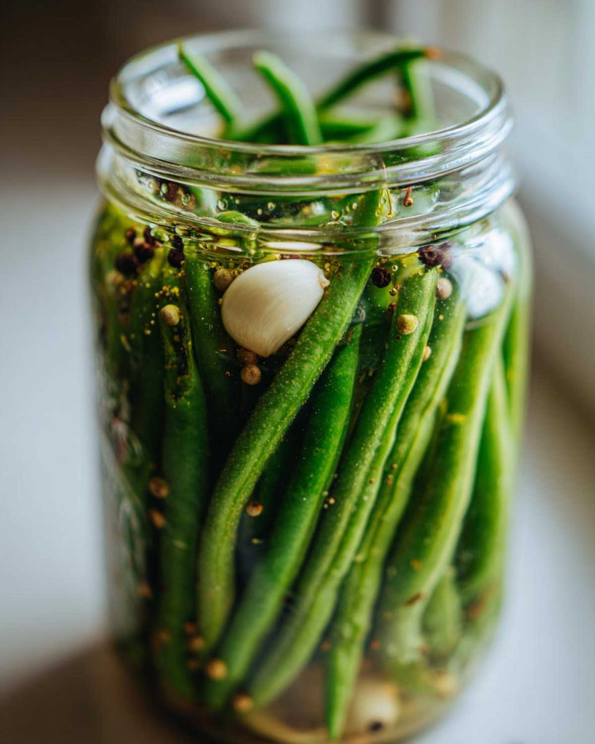 Close-up of fresh green beans packed tightly in a glass jar with a clove of garlic and peppercorns for making dilly beans.