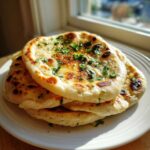 A stack of three freshly baked naan bread rounds topped with melted butter and chopped cilantro, resting on a white plate.