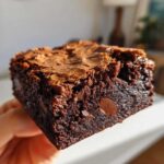 A hand holding a thick, fudgy square of brownies showing a shiny, cracked top and melted chocolate chips inside.