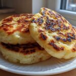 Three golden-brown, griddle-cooked irish potato farls stacked on a white plate near a sunny window.