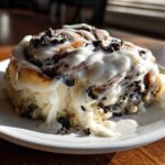 Close-up of a single, gooey oreo cinnamon rolls slice covered in white icing and chocolate cookie crumbles.