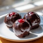 Three freshly made chocolate covered cherries with exposed bright red centers, resting on a white plate.