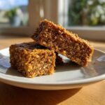 Two homemade, chewy protein bars made with oats stacked on a white plate in natural sunlight.