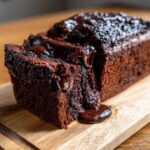 A close-up of a rich, dark hot fudge brownie bread loaf with a slice cut out, revealing gooey chocolate sauce dripping onto a wooden board.