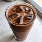 Close-up overhead view of a tall glass filled with rich, iced chocolate milk and large ice cubes.