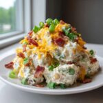A mound of creamy baked potato salad topped with shredded cheddar cheese, crispy bacon bits, and fresh green onions.
