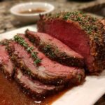 Close-up of sliced medium rare prime rib roast with herb crust, resting in jus on a white platter.