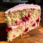 Close-up of a moist slice of cherry cake with two layers of yellow cake, cherry filling, and pink frosting.