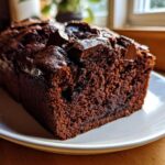 A close-up of a rich, dark chocolate banana bread loaf slice topped with melted chocolate chunks on a white plate.