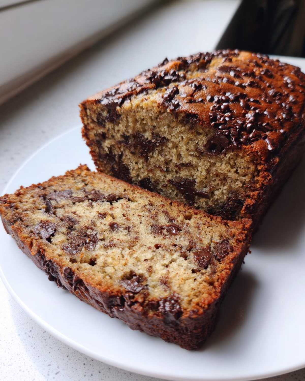 A loaf of moist chocolate chip banana bread with one slice cut, showing rich chocolate chips throughout the crumb.