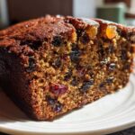 Close-up of a rich, dark slice of moist fruit cake recipe, packed with dried raisins and currants, sitting on a white plate.