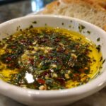 A white bowl filled with vibrant olive oil bread dip, seasoned heavily with green herbs, garlic, and red pepper flakes.