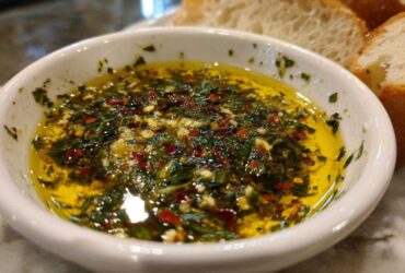 A white bowl filled with vibrant olive oil bread dip, seasoned heavily with green herbs, garlic, and red pepper flakes.