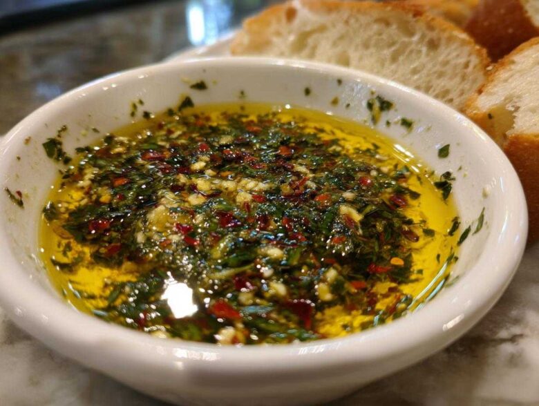 A white bowl filled with vibrant olive oil bread dip, seasoned heavily with green herbs, garlic, and red pepper flakes.