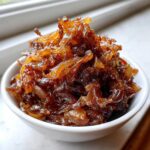 A close-up of deeply rich, glossy caramelized onions piled high in a small white bowl.