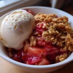 A close-up of a white bowl filled with warm rhubarb crisp topped with a scoop of vanilla ice cream.