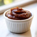 A close-up of thick, dark brown apple butter mounded in a small, white, fluted ramekin.