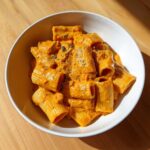A bowl of rigatoni pasta coated in a creamy, orange butternut squash pasta sauce, garnished with herbs.