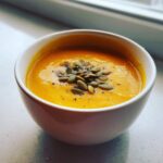 Close-up of creamy, orange roasted butternut squash soup topped with green pumpkin seeds (pepitas) and black pepper.