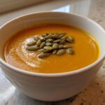 A close-up of a bowl of vibrant orange roasted butternut squash soup topped with green pumpkin seeds (pepitas).