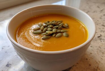 A close-up of a bowl of vibrant orange roasted butternut squash soup topped with green pumpkin seeds (pepitas).