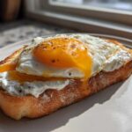 A thick slice of toasted bread topped with creamy ricotta cheese and a runny sunny-side-up egg, seasoned with pepper. This is a savory ricotta toast.