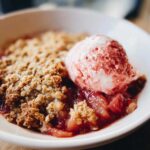 Close-up of a warm serving of rhubarb crisp topped with a scoop of strawberry swirl ice cream.