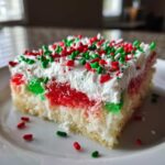A festive slice of christmas poke cake featuring layers of white cake, red and green gelatin, whipped topping, and holiday sprinkles.