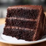 Close-up of a moist, three-layer slice of dark chocolate cake recipe covered in thick chocolate frosting.