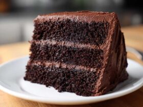 Close-up of a moist, three-layer slice of dark chocolate cake recipe covered in thick chocolate frosting.
