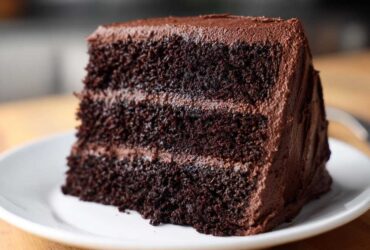 Close-up of a moist, three-layer slice of dark chocolate cake recipe covered in thick chocolate frosting.