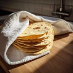 A warm stack of freshly made corn tortillas wrapped partially in a white cloth on a wooden surface.