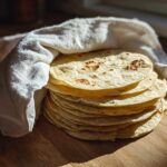 A warm stack of freshly made corn tortillas wrapped partially in a white cloth on a wooden surface.