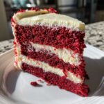 A tall, moist slice of red velvet cheesecake showing three layers of deep red cake alternating with thick layers of white cream cheese frosting.