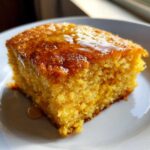 A close-up of a square slice of moist, golden sweet potato cornbread drizzled with honey on a white plate.