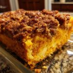 A close-up view of a square serving of bright orange sweet potato souffle topped with a dark brown, sugary crumb topping.