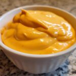 A close-up of thick, vibrant orange nacho cheese sauce in a small white bowl.