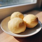 Three smooth, golden balls of freshly made fufu served on a light ceramic plate near a window.