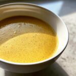 Close-up of creamy, vibrant yellow cauliflower soup with fine bubbles on the surface in a white bowl.