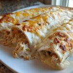 Three rolled white chicken enchiladas covered in creamy sauce and browned cheese on a white plate.