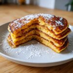 A stack of four golden brown almond flour pancakes dusted with powdered sugar and drizzled with syrup.