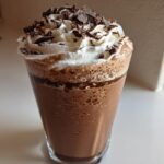 A tall glass filled with blended frozen hot chocolate, topped with whipped cream and chocolate shavings.