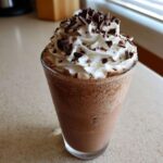 A tall glass filled with blended frozen hot chocolate topped with whipped cream and chocolate shavings.