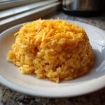 A mound of creamy, orange-colored cheesy rice topped with shredded cheddar cheese on a white plate.