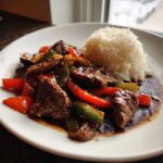 A close-up of a plate featuring savory pepper steak mixed with colorful bell peppers and a side of fluffy white rice.