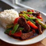 A plate featuring savory pepper steak stir-fry with red peppers and green onions, served alongside a mound of white rice.