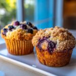 Two golden baked blueberry muffins, one with a streusel topping, sitting on a white plate.