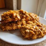 Close-up of four moist, orange-hued breakfast cookie oat treats stacked and resting on a white plate.
