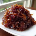 A close-up of deeply browned, glossy caramelized onions piled high on a white square plate.
